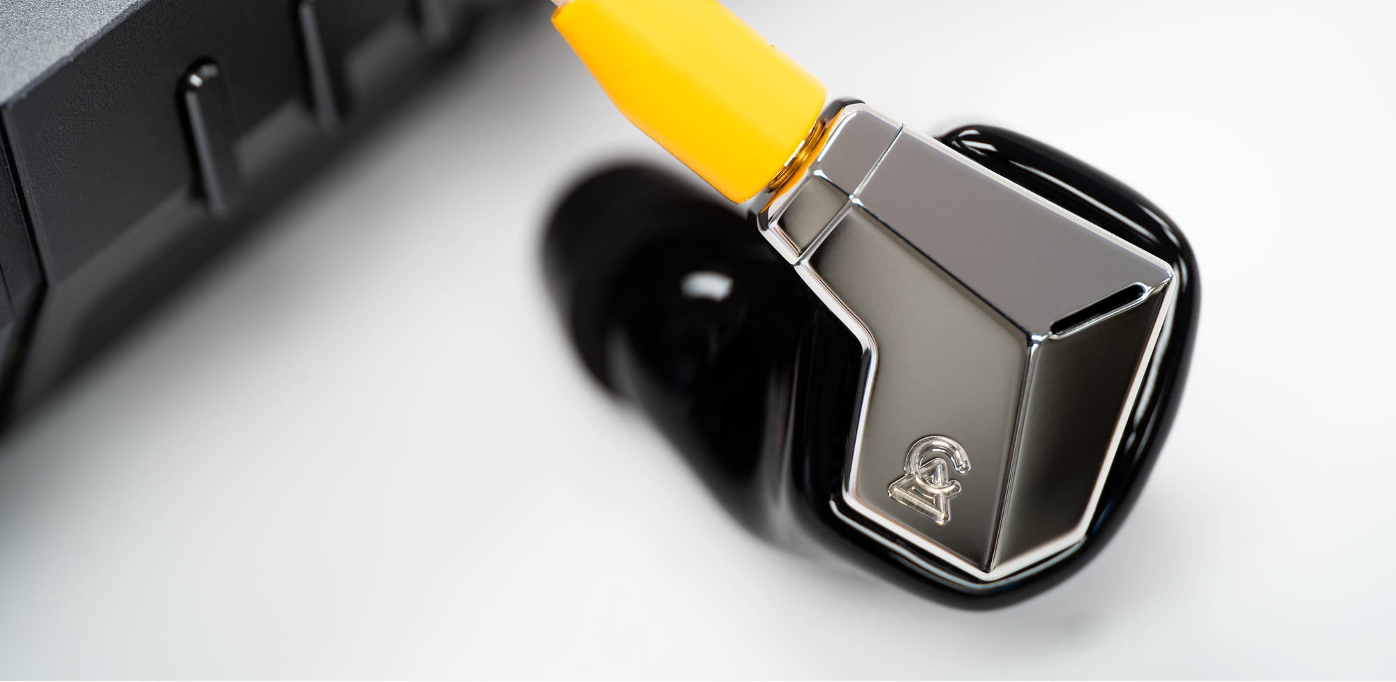 A close-up of a black and silver in-ear monitor earpiece with a bright yellow connector, resting next to a dark gray electronic device on a white surface.